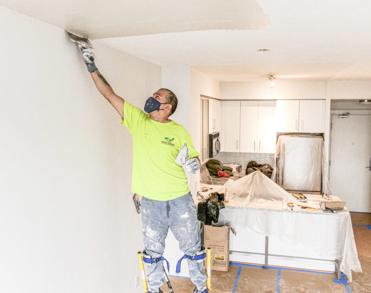 Professional house painter wearing mask and working on popcorn ceiling removal using stilts, popcorn ceiling removal, how to remove popcorn ceiling, popcorn ceiling, how to paint popcorn ceiling, painted popcorn ceiling removal, popcorn paint removal